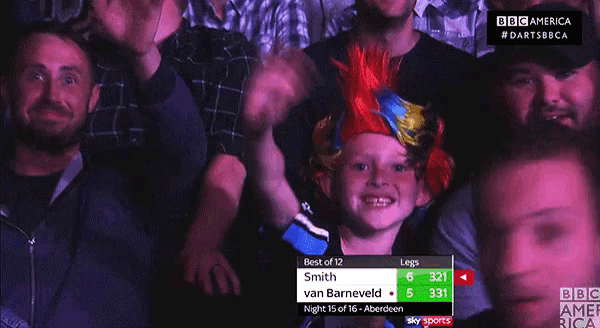 a young boy wearing a colorful mohawk stands in a crowd of people watching a game of darts