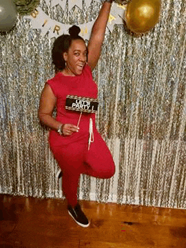 a woman in a red jumpsuit is standing on one leg in front of a silver curtain and holding a sign .