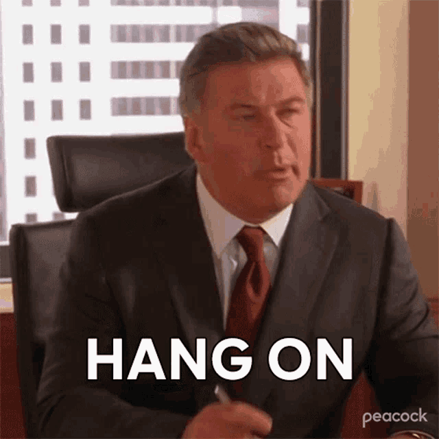 a man in a suit and tie is sitting at a desk with the words hang on written on his face .