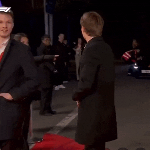 a man in a suit and coat is standing on a red carpet while people point at him .