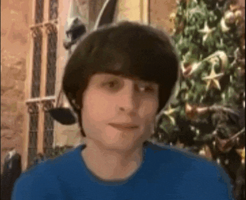 a young man in a blue shirt is standing in front of a christmas tree and smiling .