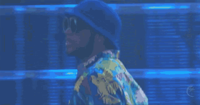 a man wearing a hat and sunglasses stands in front of a blue background