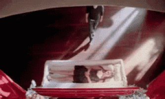 a woman is laying in a glass coffin with a man walking behind her .