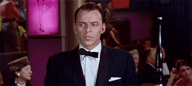a man in a tuxedo and bow tie stands in front of a crowd of people