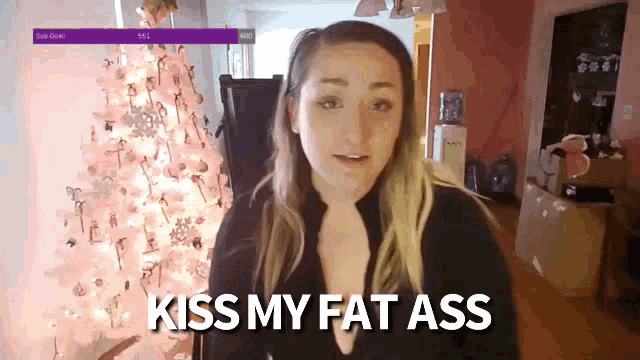a woman is standing in front of a pink christmas tree and says kiss my fat ass
