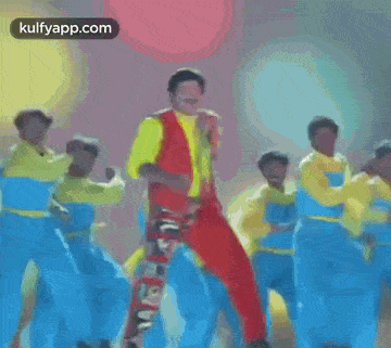 a man in a yellow and red vest is dancing with a group of people in blue pants .