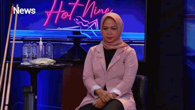 a woman wearing a hijab sits in front of a neon sign that says hot nine