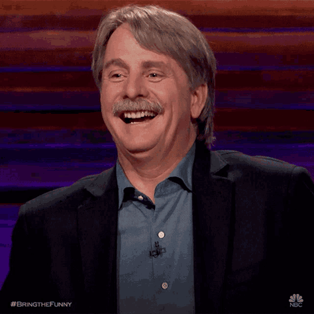 a man with a mustache is laughing while wearing a suit and blue shirt