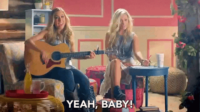 a woman playing a guitar next to another woman who is sitting on a couch and saying yeah baby