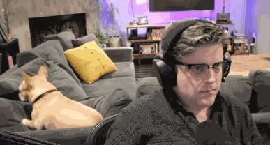a man wearing headphones and glasses sits on a couch with a dog behind him
