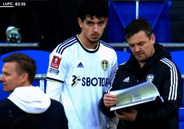 a soccer player wearing a sbotop jersey stands next to two men