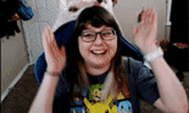 a woman wearing glasses and a pikachu shirt is smiling and waving her hands .