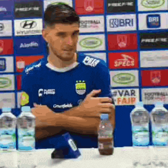 a man in a blue jersey is sitting at a table with bottles of water in front of him .