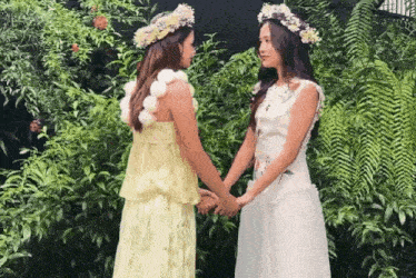 two women wearing flower crowns hold hands in a garden