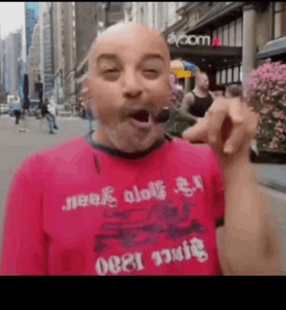 a bald man wearing a red shirt that says ' neak ololi ' on it is standing on a city street