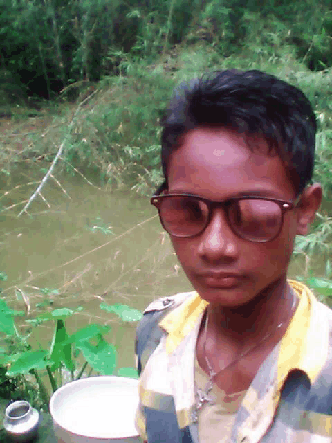 a young boy wearing glasses and a plaid shirt is standing in front of a body of water