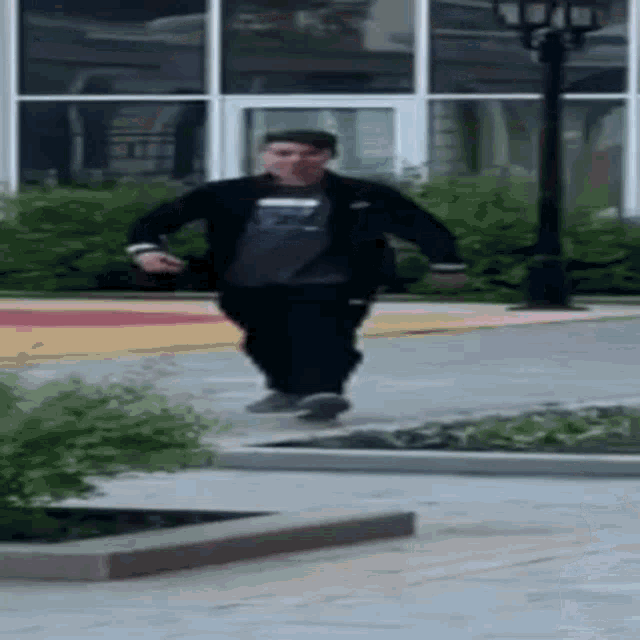 a man is walking down a sidewalk in front of a building while wearing a black jacket .