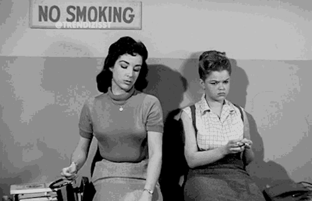 two women are sitting next to each other in front of a sign that says no smoking .