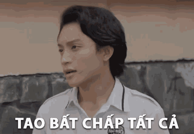a young man in a white shirt is standing in front of a wall with the words tao bat chap tat ca above him