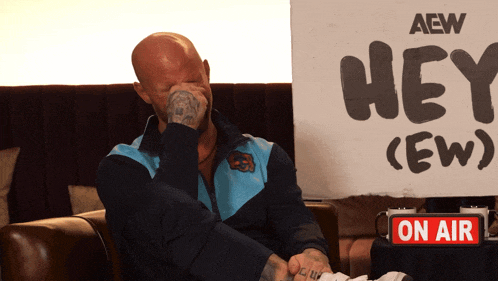 a bald man sits in front of a sign that says hey ( ew )