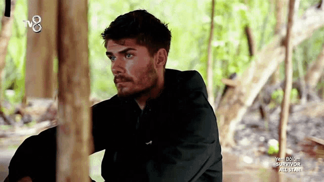 a man in a black hoodie sits in the woods with a tv8 logo in the background