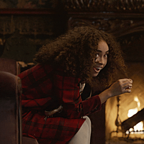 a woman with curly hair is sitting in front of a fireplace smiling