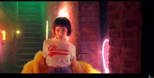 a woman wearing a white t-shirt with the word glamour on it is standing in front of a neon sign .