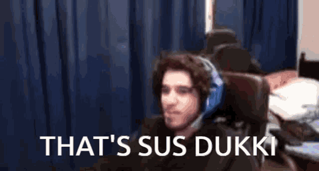 a man wearing headphones is sitting in front of a blue curtain with the words `` that 's sus dukki '' .