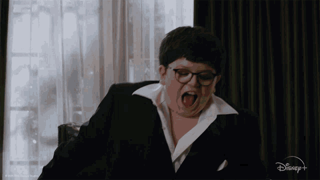 a boy with glasses and a black suit is sitting in a chair with his mouth open and a disney logo behind him