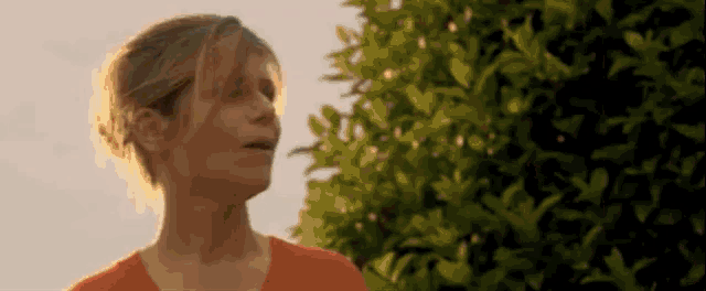 a woman in an orange shirt is standing in front of a tree with leaves .