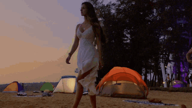 a woman in a white dress is walking on a beach near tents