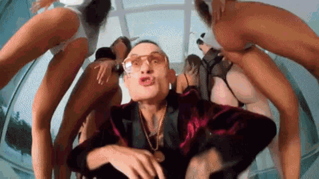 a man in a suit and sunglasses is surrounded by naked women in bikinis .