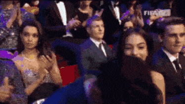 a group of people are sitting in a theatre applauding a man in a suit and tie .
