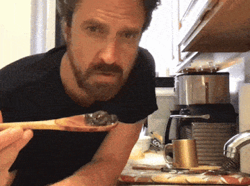 a man with a beard is holding a wooden spoon in front of a coffee maker