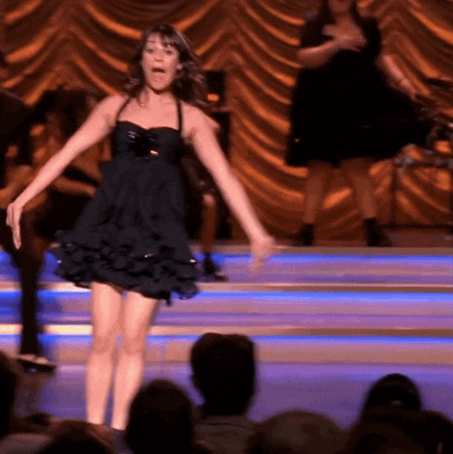 a woman in a black dress is dancing on stage in front of a crowd