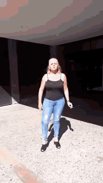 a woman in a black tank top and blue jeans is standing on a sidewalk in front of a building .