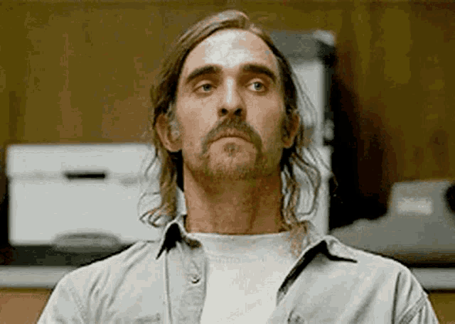 a man with long hair and a beard is standing in a kitchen looking at the camera .