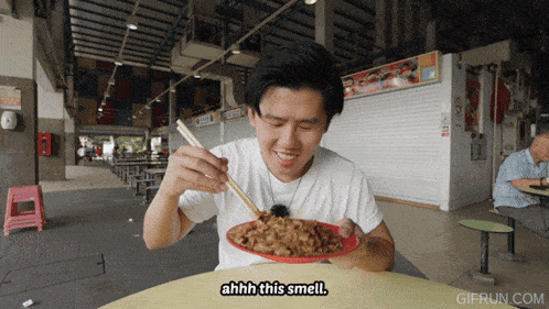 a man sitting at a table eating a plate of food with chopsticks and the words ahhh this smell below him