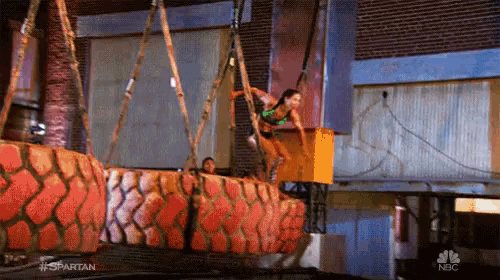 a woman in a green tank top is hanging from a crane on a nbc show called spartan .