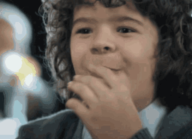 a young boy with curly hair is smiling and covering his mouth with his hand