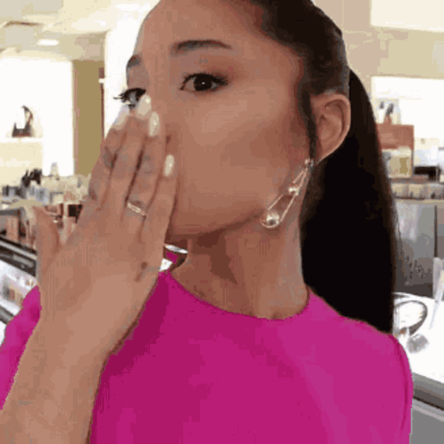 a woman wearing a pink shirt and clear earrings covering her mouth with her hand