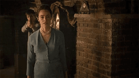 a woman in a blue dress and pearls is walking in front of a brick wall .