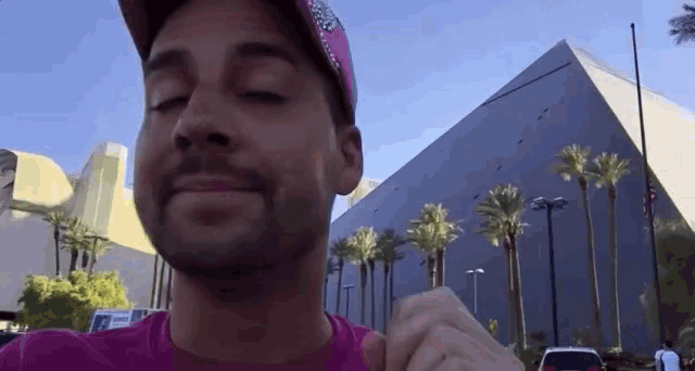 a man wearing a pink shirt and a pink hat is standing in front of a building with palm trees .