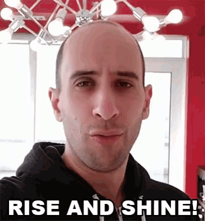 a bald man is taking a selfie in front of a chandelier and the words rise and shine .