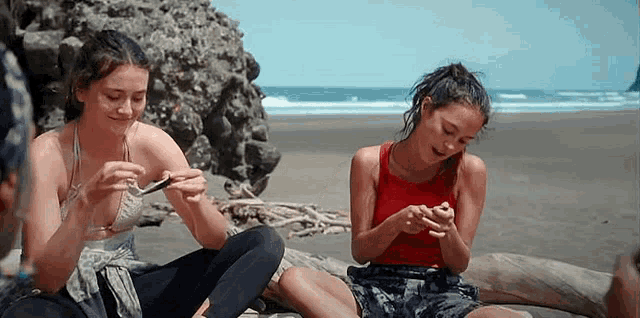 two young women are sitting on a beach looking at their phones .