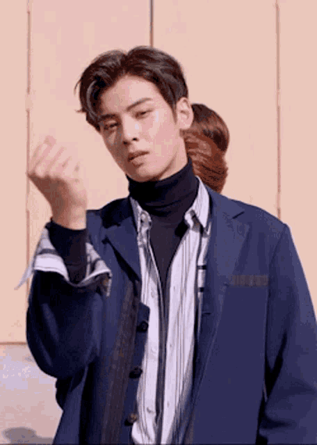 a young man wearing a blue jacket and a turtleneck is making a heart shape with his hand