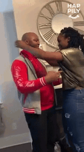 a man in a red jacket is being hugged by a woman in front of a clock .