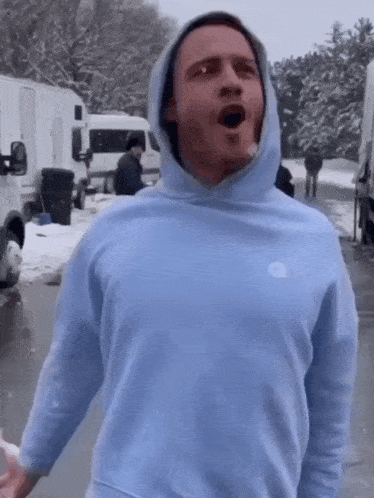 a man wearing a light blue hoodie is standing in the snow with his mouth open .