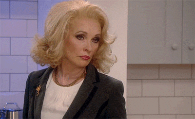 a woman with blonde hair and a gray jacket is standing in a kitchen looking at the camera .