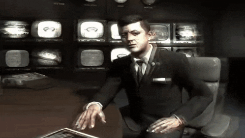 a man in a suit and tie sits at a desk in front of a wall of monitors with the number 97 on them